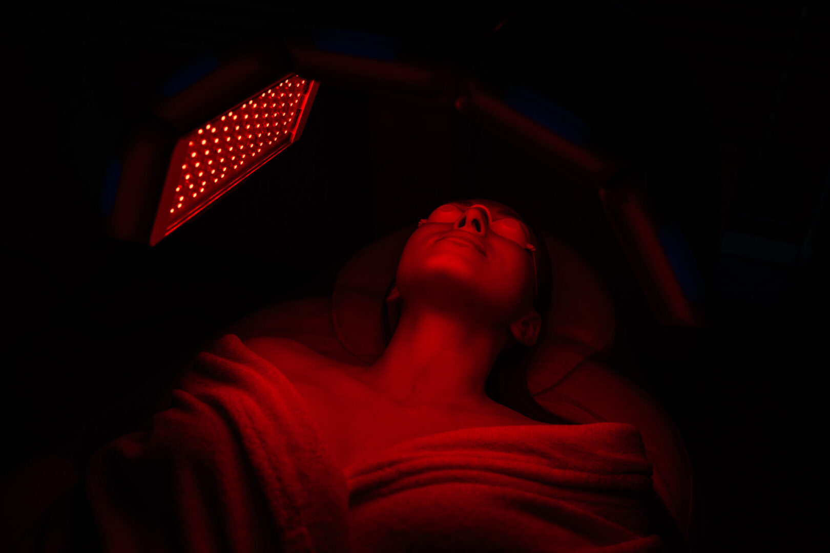 Top view of relaxed female client having red LED light facial photodynamic therapy treatment in modern beauty salon. Young woman in protective glasses patient LED Skin Care Spa