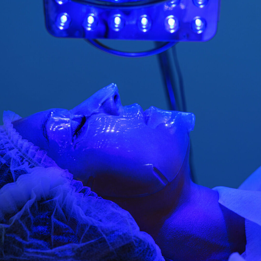 Young woman having blue LED light facial therapy treatment in beauty salon. Beautician wearing face mask maintaining safety procedures during appointment. Beauty, new normal and wellness LED Skin Care Spa